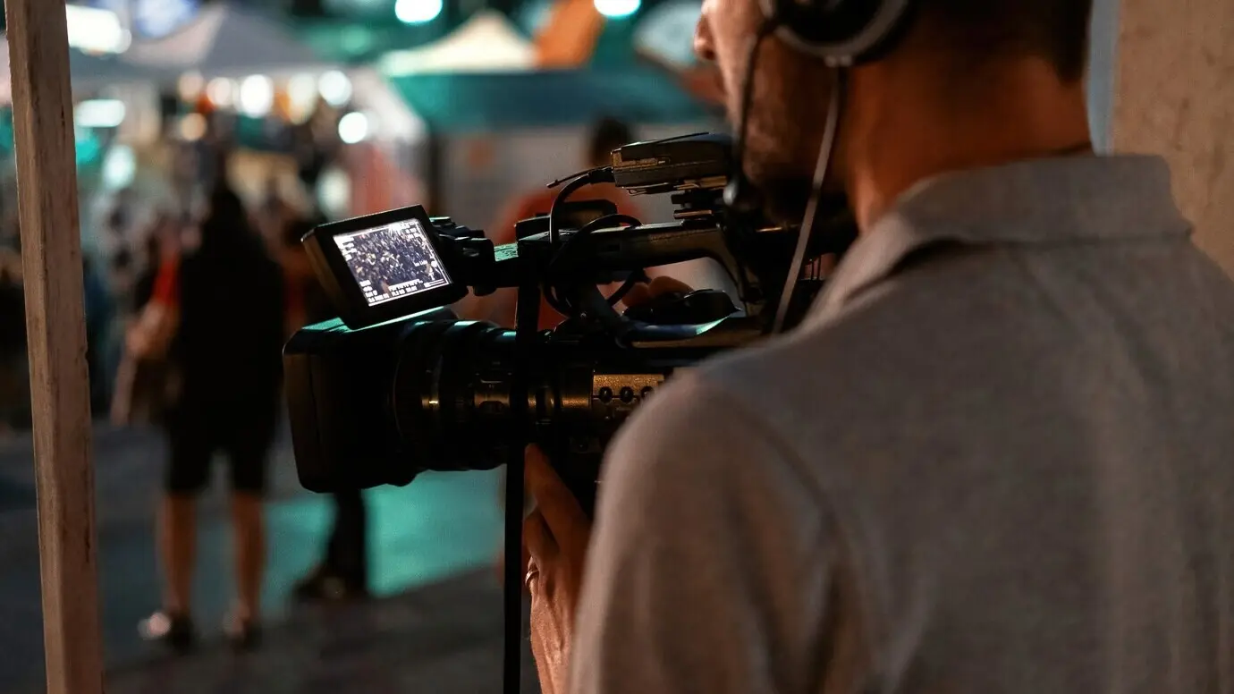 A professional camera operator filming people watching football in a public place at night.