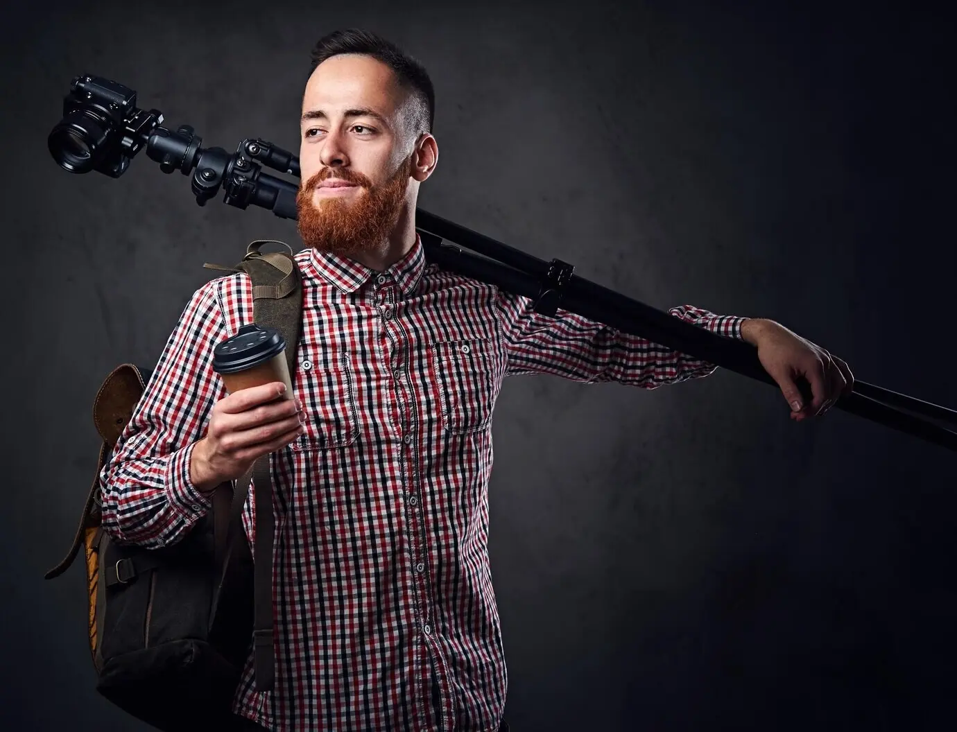 A red-haired, bearded freelance photographer with a tripod and a backpack.